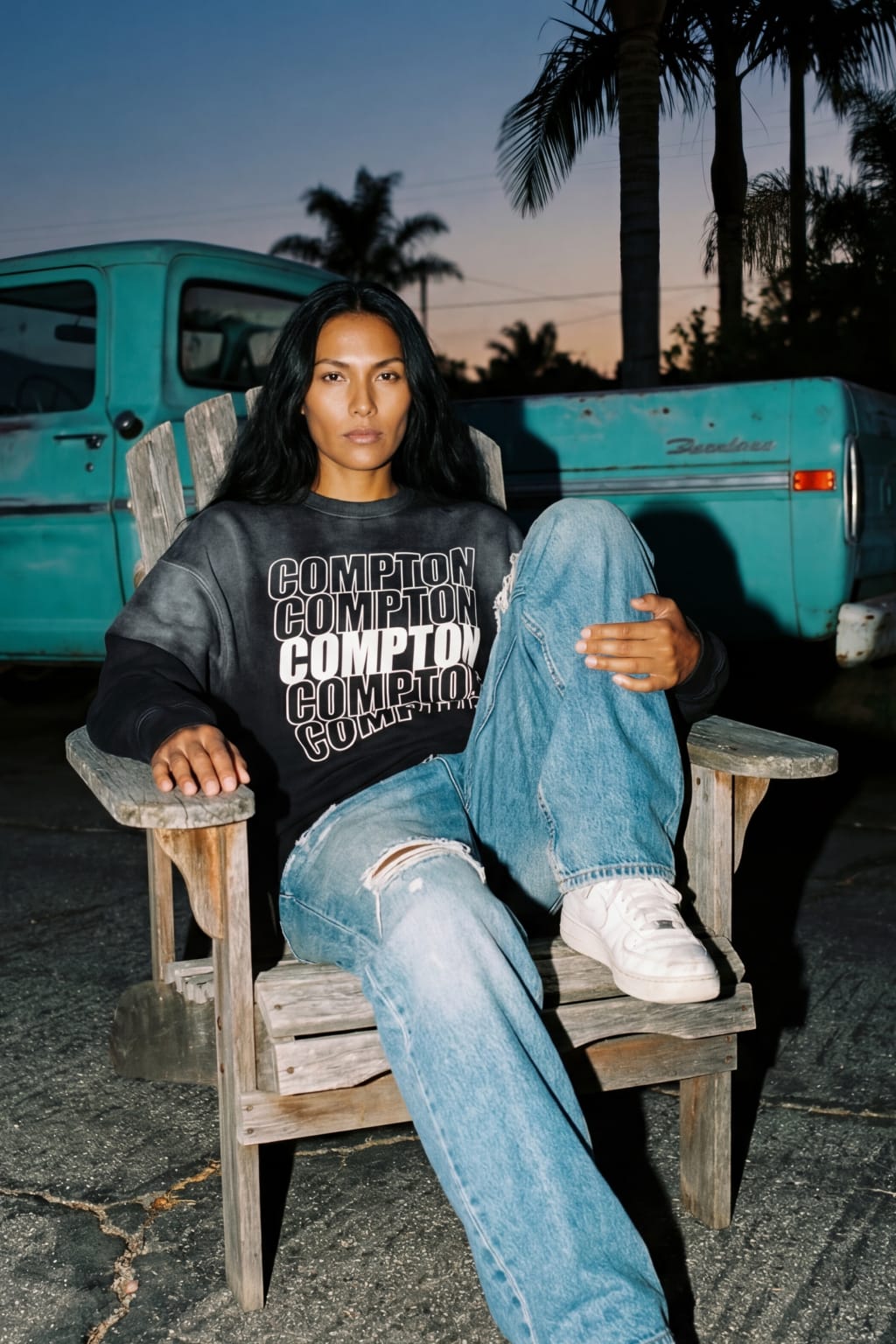 Female model wearing black Sunfade Compton sweatshirt in relaxed fit, seated outdoors with vintage truck in California streetwear editorial scene