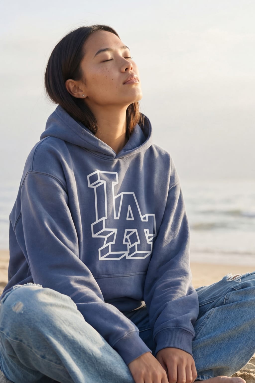 Woman in blue LA hoodie sitting on beach with eyes closed in sunset light