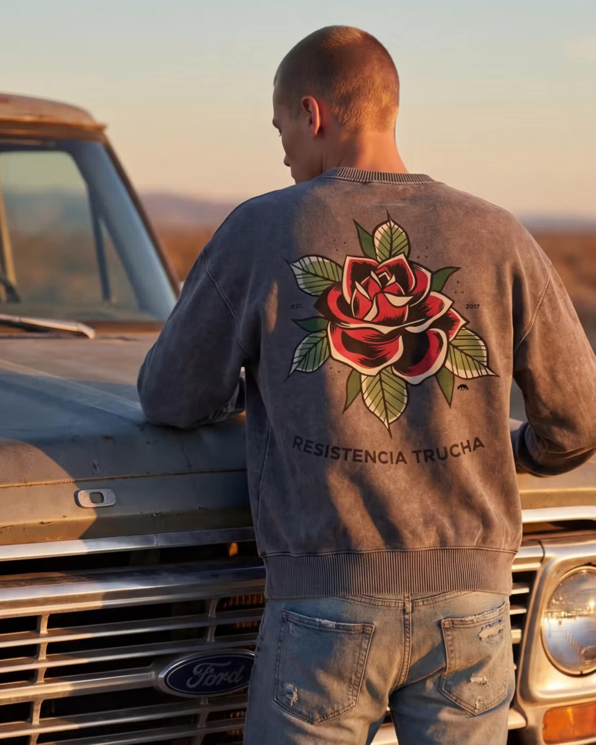 Model wearing the FERN + FIN RESISTENCIA TRUCHA mineral wash sweater beside a vintage truck in warm desert light.